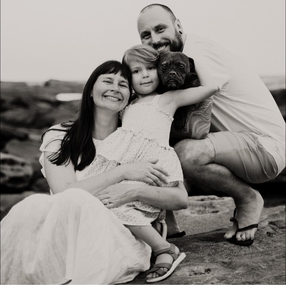 A smiling family of three, including a young girl holding a small dog, sits on rocks by the water. The photo is in black and white, and all appear happy and close together.