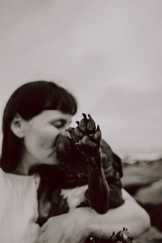 A woman kisses her dog while holding it close; the dog's paw reaches toward the camera, creating a blurred, affectionate pet photography scene in black and white.