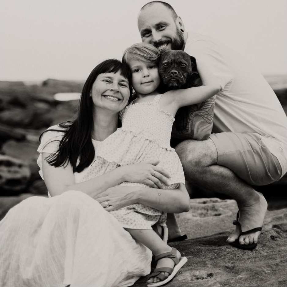 A happy family poses on rocky ground: a woman in a long dress, a young girl in a polka-dot dress, and a smiling man crouching beside them. The girl holds a small dog, capturing the joy of pet photography as they all smile warmly at the camera.