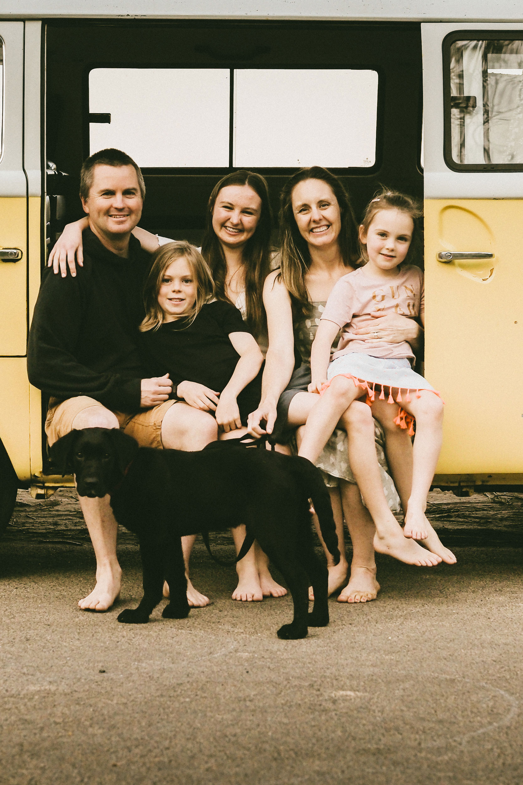 A barefoot family of four and a black dog sit and smile together in front of a yellow and white van, capturing joyful moments from their photo sessions on the street.
