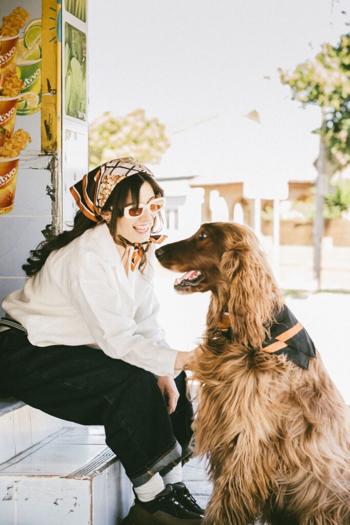 A woman wearing sunglasses and a patterned headscarf smiles while sitting on steps next to a large, long-haired dog. They appear happy and are outdoors near an ice cream shop.