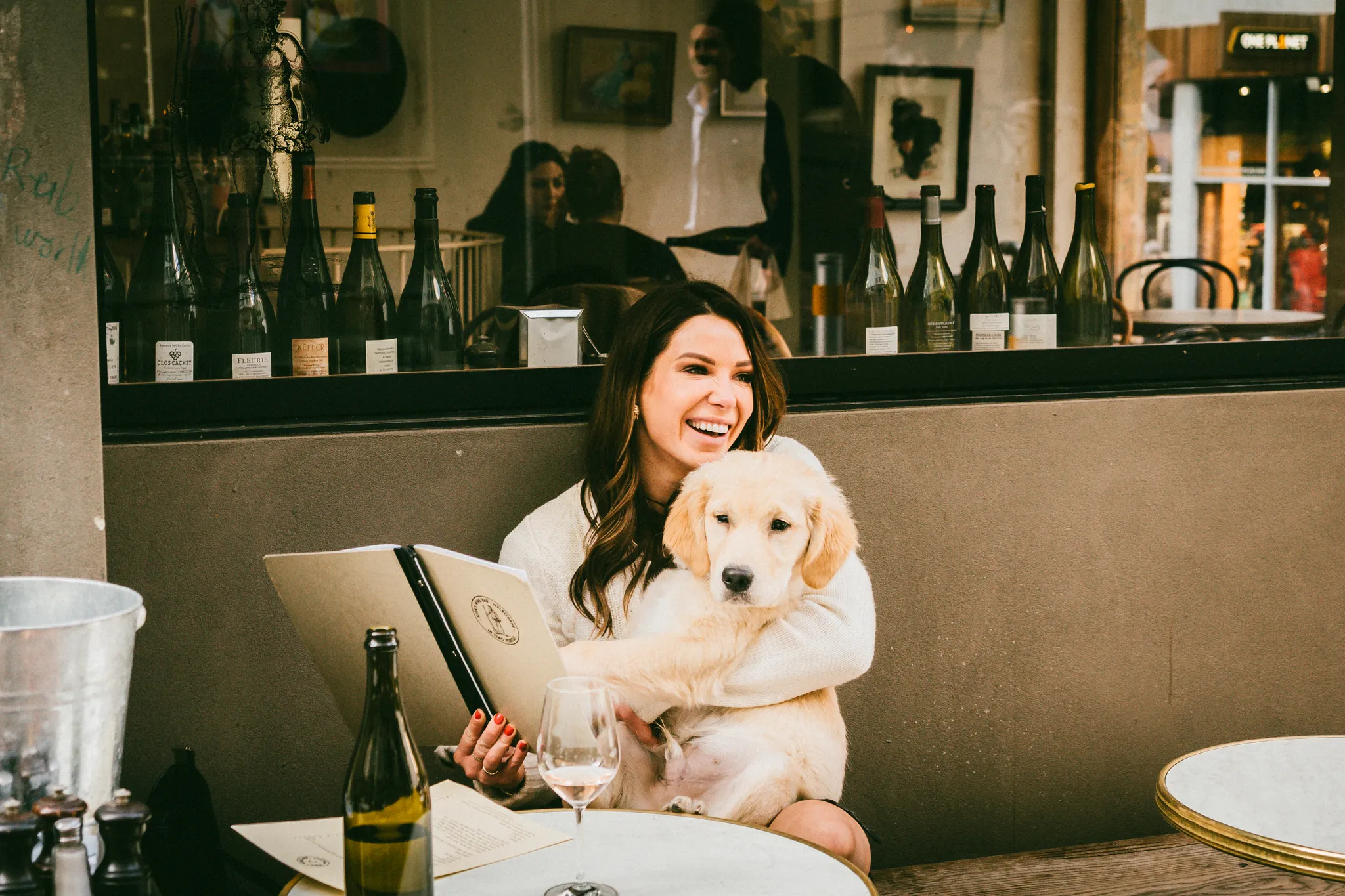 A woman sits at an outdoor café table, smiling and holding a Golden Retriever puppy for a charming dog photography moment. She has a menu in hand, with wine bottles behind her and a glass of wine on the table.