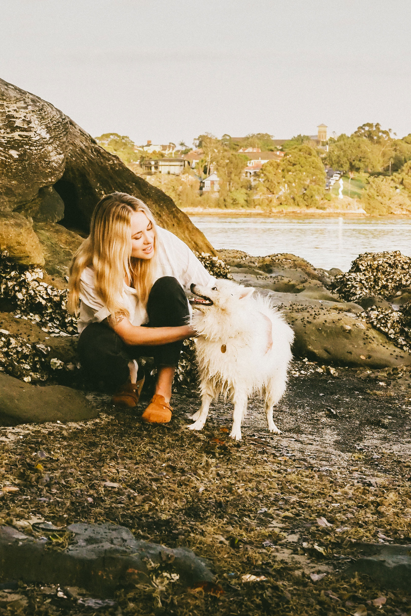 A woman with long blonde hair smiles and pets a fluffy white dog on a rocky shore, with green trees and houses visible across the water in the background.