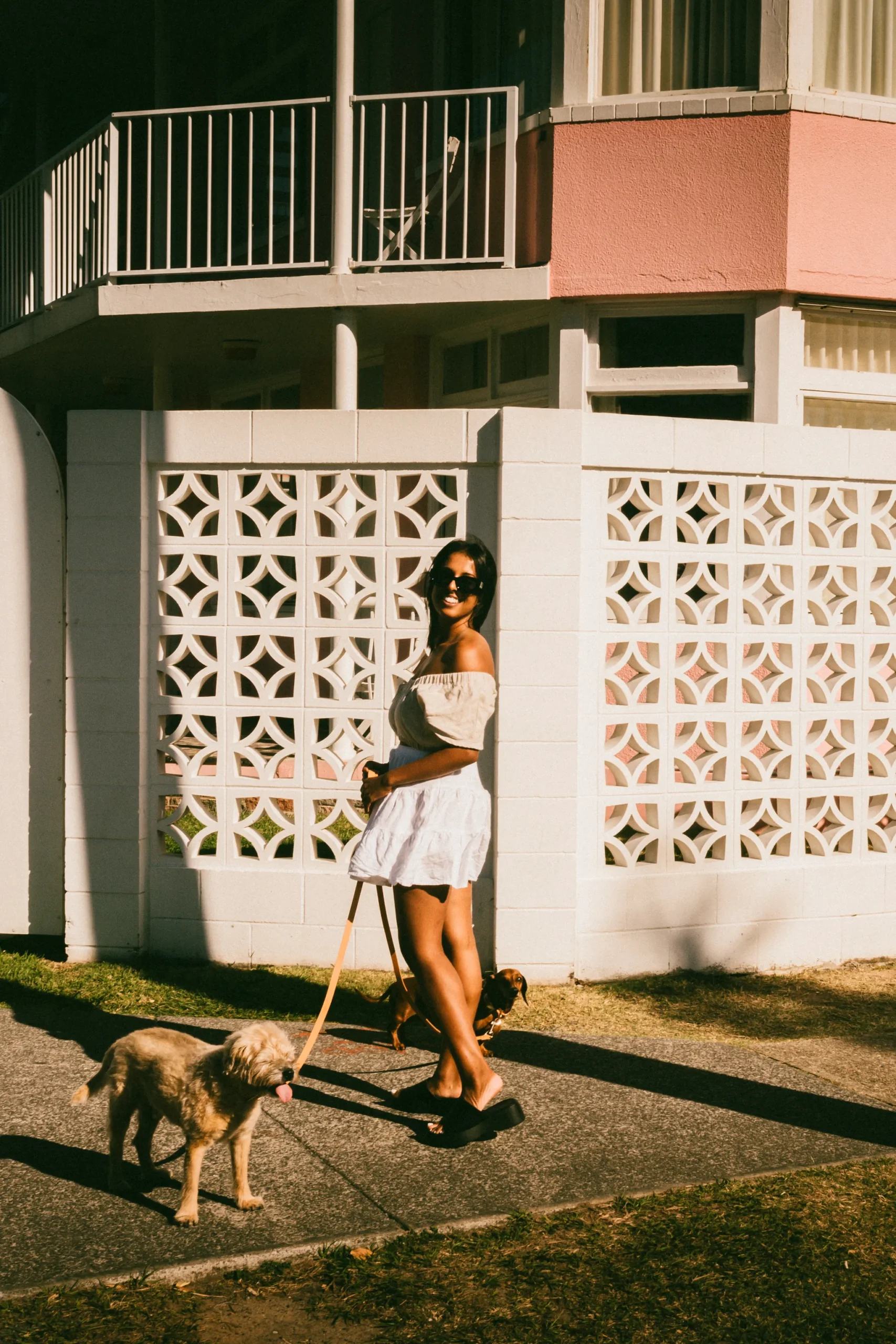 A woman in sunglasses and a white dress walks two dogs on a sunny sidewalk, showcasing stylish dog photography in front of a modern building with a decorative white wall and pink accents.