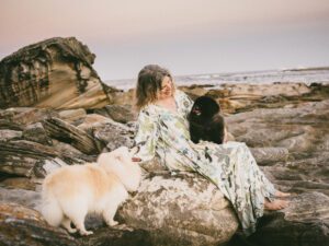 A woman sitting on the rocks with her 16 year old and 6 year dogs, with the sun setting in the background. 